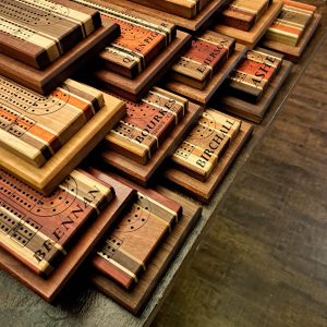 12 Handmade 2-tier cribbage boards from Canada, some personalized, stacked on a harvest table by Bergeron Woodgrains.